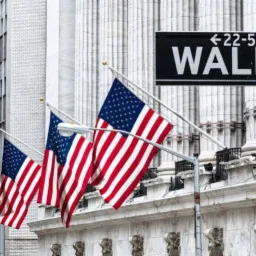 Trzy amerykańskie flagi powiewają przed budynkiem z kolumnami i tabliczką z napisem Wall St.
