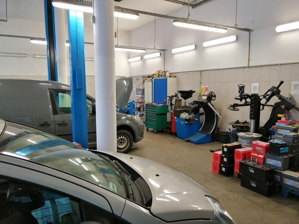 Wnętrze warsztatu samochodowego z podnośnikiem, wyważarką do kół i dwoma samochodami, jeden z otwartą maską. Na pierwszym planie fragment srebrnego samochodu.