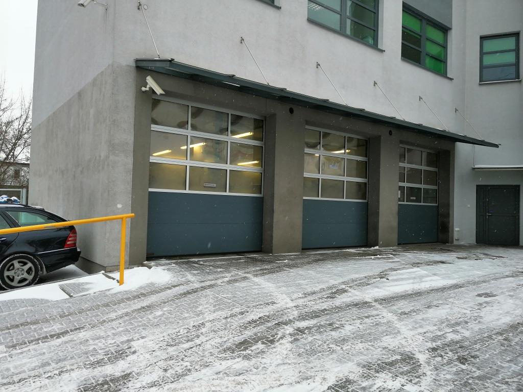Zewnętrzny widok warsztatu samochodowego z trzema bramami garażowymi w zimowej scenerii, częściowo zasypanymi śniegiem. Czarny samochód osobowy zaparkowany z boku budynku, widoczny fragment żółtej...