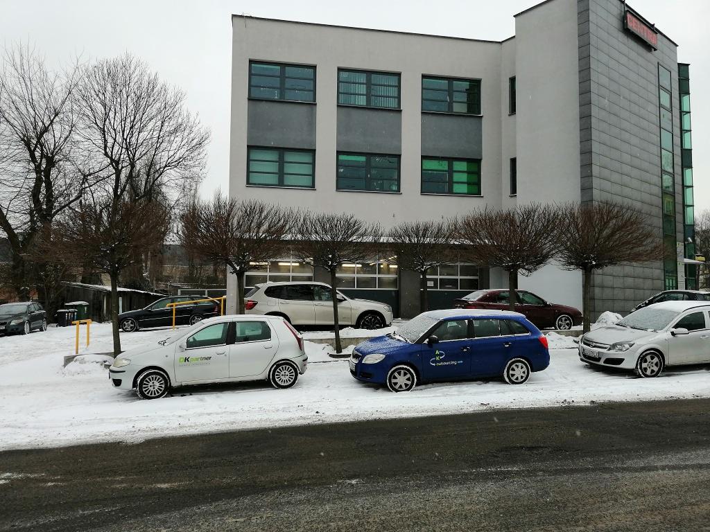 Zimowy widok na budynek warsztatu samochodowego z zaparkowanymi, lekko ośnieżonymi autami przed wejściem.