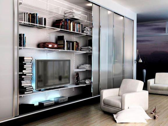 Biała, otwarta garderoba z przesuwnymi drzwiami, wyposażona w półki z książkami, dekoracjami i telewizorem, oświetlona od dołu, z dwoma fotelami w tle.