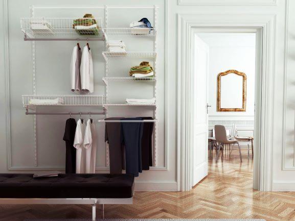 Biały, modułowy system przechowywania ubrań z koszami i półkami, zamontowany na ścianie w jasnym wnętrzu z parkietem i otwartymi drzwiami do kolejnego pokoju z lustrem.