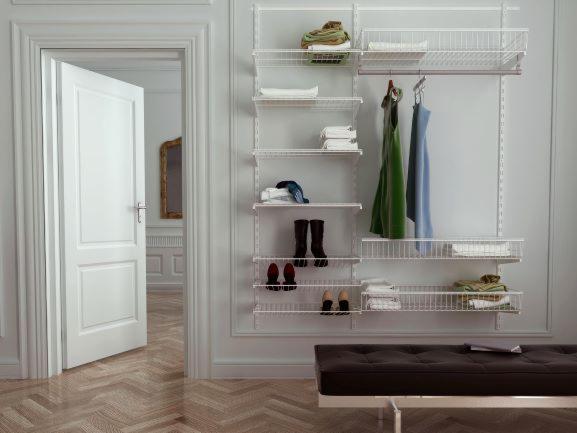 Biały, modułowy system przechowywania z metalowymi półkami i koszami, z ubraniami, butami i złożonymi ręcznikami, ustawiony w pomieszczeniu z otwartymi drzwiami i parkietem w jodełkę.