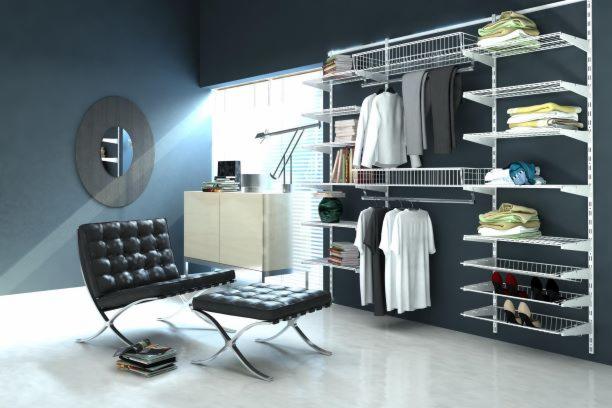 System przechowywania z białymi, metalowymi półkami i koszami zamontowany na ścianie, wypełniony ubraniami, butami i książkami, w tle widoczne krzesło Barcelona i szafka.