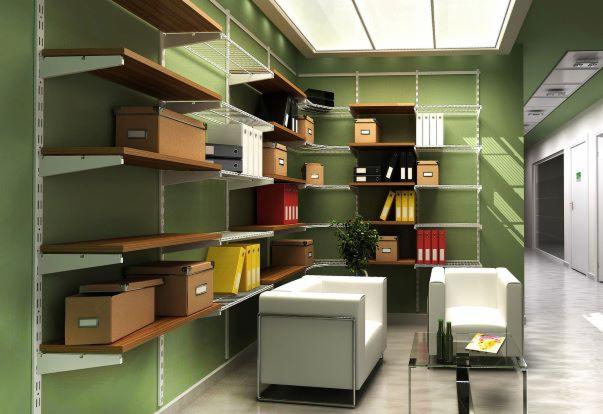 System regałów z drewnianymi półkami i metalowymi wspornikami na zielonej ścianie, wypełniony pudełkami, segregatorami i dokumentami, z białymi fotelami i szklanym stolikiem w minimalistycznym biurze.