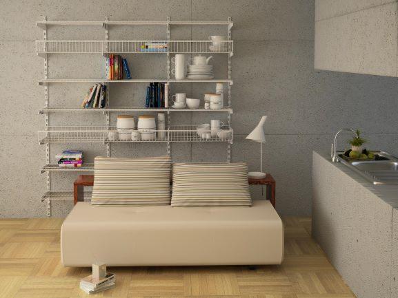 System modułowych, metalowych półek na ścianie z książkami, naczyniami i dekoracjami, ustawiony za beżową sofą z poduszkami w paski, w tle widoczny fragment blatu kuchennego z umywalką.