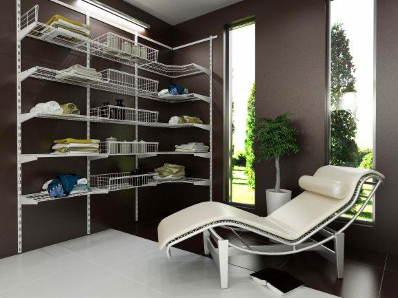 System regałów drucianych w szafie wnękowej z białymi półkami i koszami, uporządkowane ubrania i akcesoria, w tle minimalistyczny leżak i okno z widokiem na zieleń.