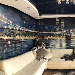 Like Denis Shapel - Panoramiczny widok salonu z ręcznie malowanym muralem przedstawiającym nocny krajobraz miejski z mostem, odbijającym się w wodzie, oraz kremową sofą narożną i błyszczącym sufitem.