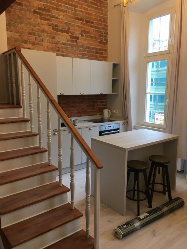 Jasna kuchnia z ceglaną ścianą w tle, widoczne schody z drewnianymi stopniami i białą balustradą, wyspa kuchenna z dwoma stołkami barowymi oraz zwinięty dywan na podłodze.