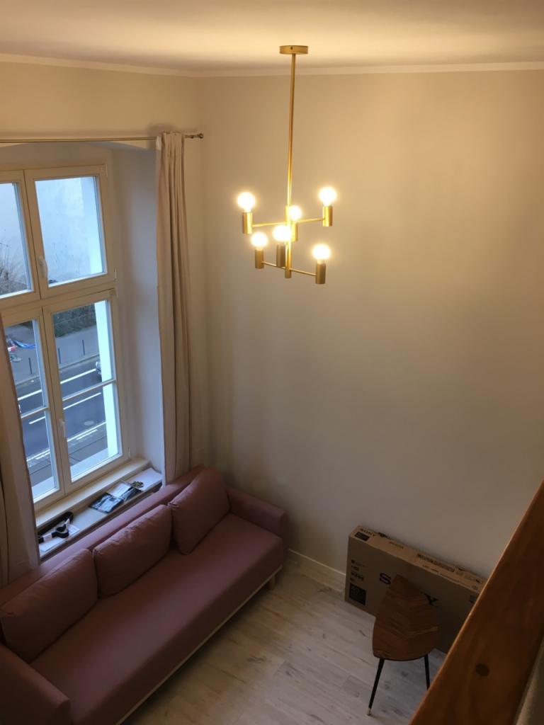 Nowoczesny salon z różową sofą pod oknem, złoty żyrandol z żarówkami kulkami i drewnianą podłogą, widok z góry.