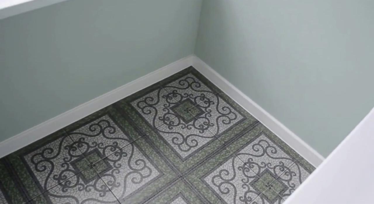 Fragment wykończonego wnętrza z dekoracyjną, wzorzystą podłogą z płytek w odcieniach szarości i zieleni, jasne ściany i biała listwa przypodłogowa.