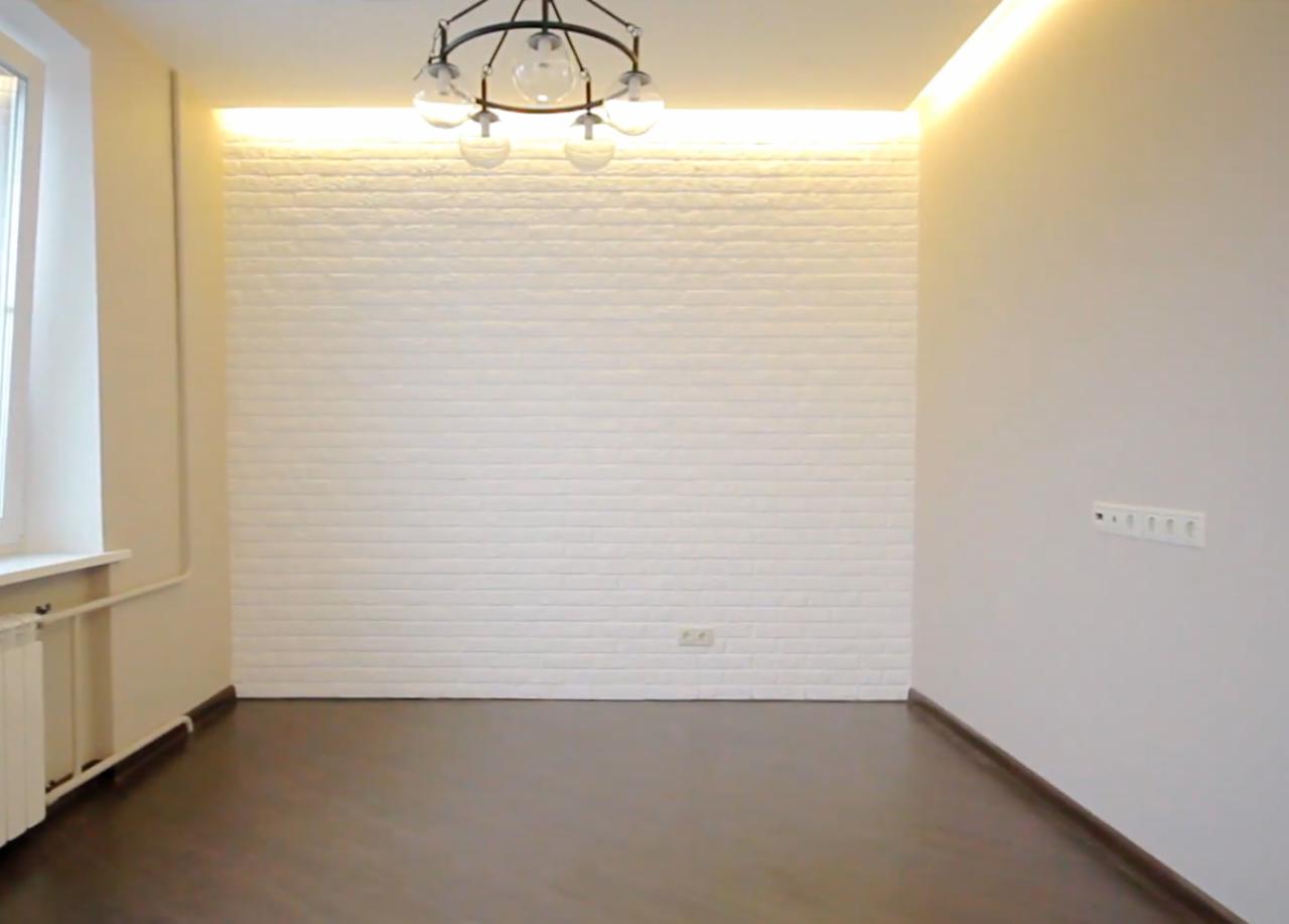 Jasne, puste wnętrze z białą ścianą wykończoną dekoracyjną cegłą, ciemną podłogą, nowoczesnym oświetleniem sufitowym i grzejnikiem pod oknem.