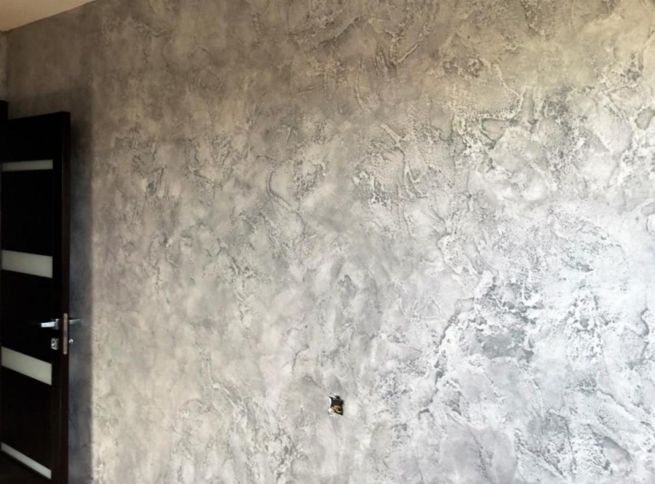 Ściana w odcieniach szarości z dekoracyjną fakturą imitującą marmur, widoczny fragment czarnych drzwi z białymi paskami po lewej stronie oraz gniazdko elektryczne na ścianie.