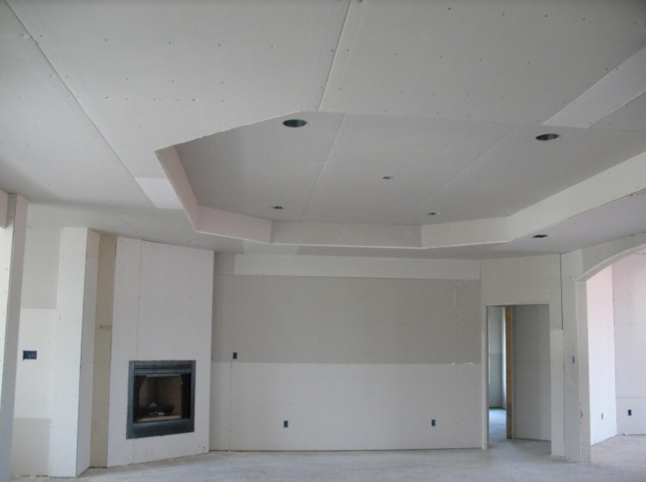 Wnętrze w trakcie wykańczania, widoczne ściany i sufit pokryte płytami kartonowo-gipsowymi, przygotowane do dalszych prac; wbudowany kominek z czarną obudową, brak wykończenia podłogi, sufit...