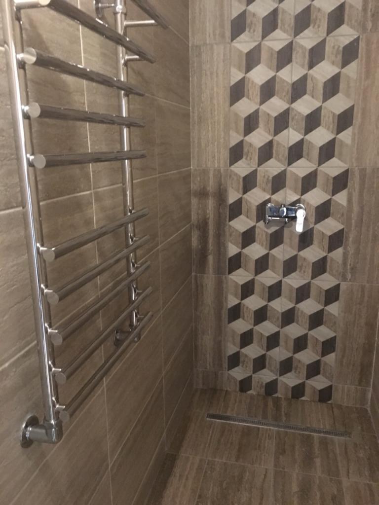 Wykończona łazienka z grzejnikiem drabinkowym, brązowymi płytkami imitującymi kamień i dekoracyjnym panelem z płytek z motywem trójwymiarowych kostek nad chromowaną baterią prysznicową.