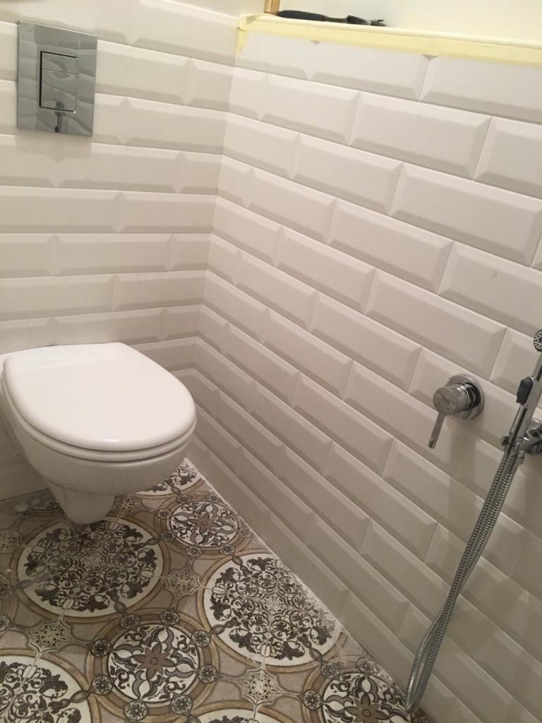 Wykończona łazienka z białą podwieszaną toaletą, wzorzystą podłogą i białą cegiełkową glazurą z chromowaną armaturą.