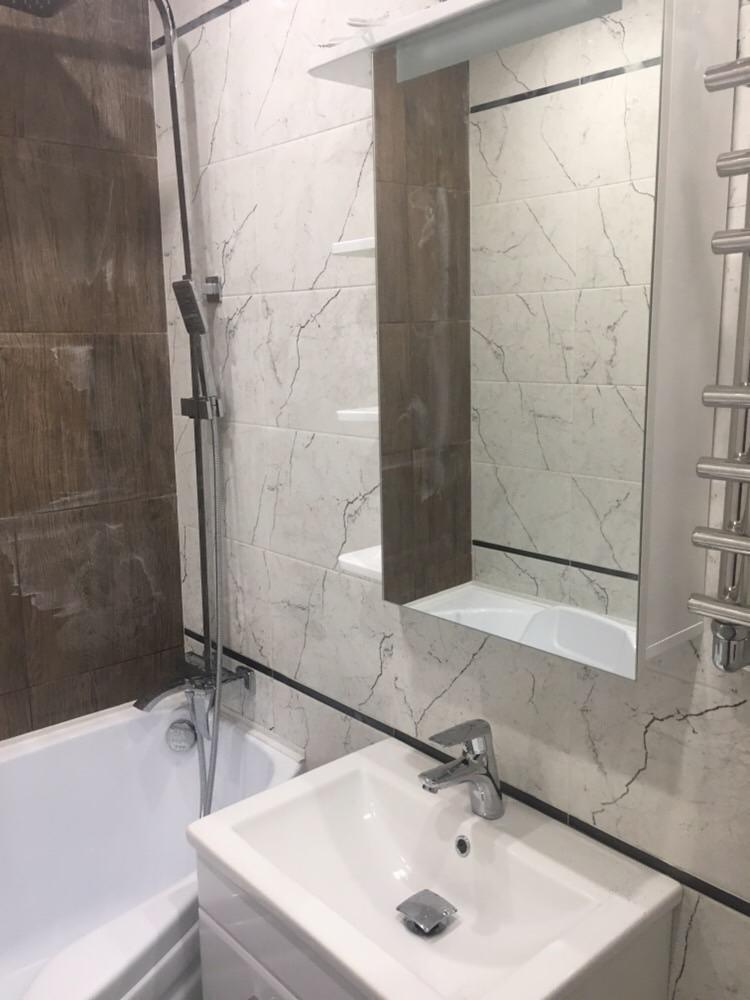 Umywalka z chromowaną baterią i odpływem w wykończonej łazience z marmurowymi płytkami i brązowymi akcentami, widoczny fragment wanny i lustra.