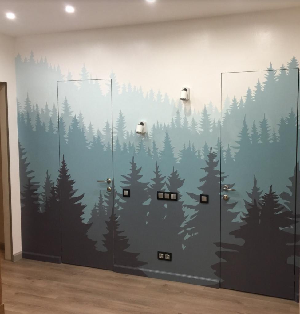 Wnętrze z pomalowaną ścianą i drzwiami w motyw lasu iglastego, z widocznymi włącznikami światła i lampkami ściennymi.