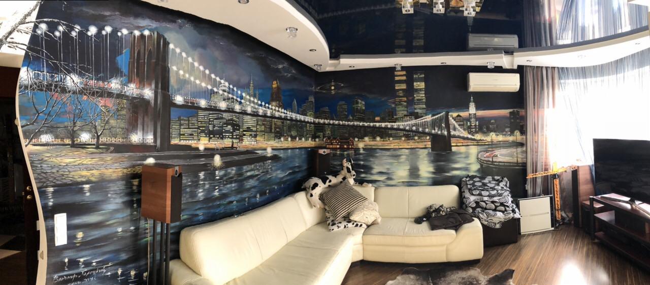 Panoramiczny widok salonu z ręcznie malowanym muralem przedstawiającym nocny krajobraz miejski z mostem, odbijającym się w wodzie, oraz kremową sofą narożną i błyszczącym sufitem.