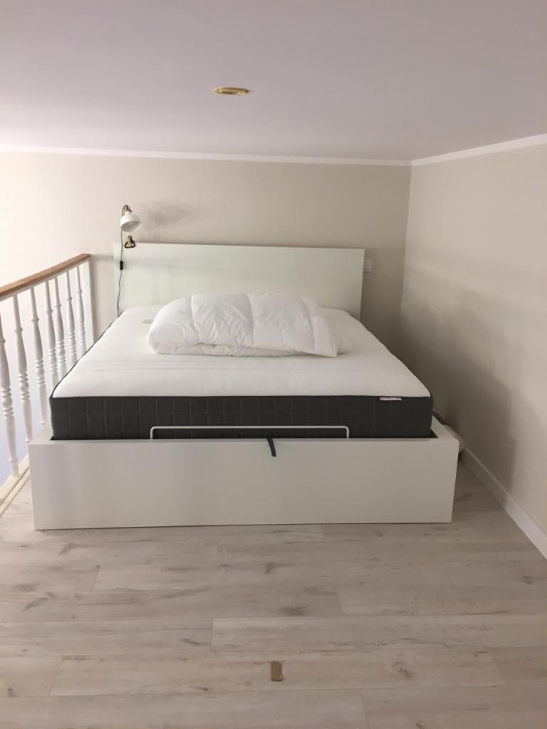 Biała sypialnia na antresoli: łóżko z szarą ramą materaca i białą pościelą pod lampką ścienną, widok na drewnianą podłogę i białe barierki.