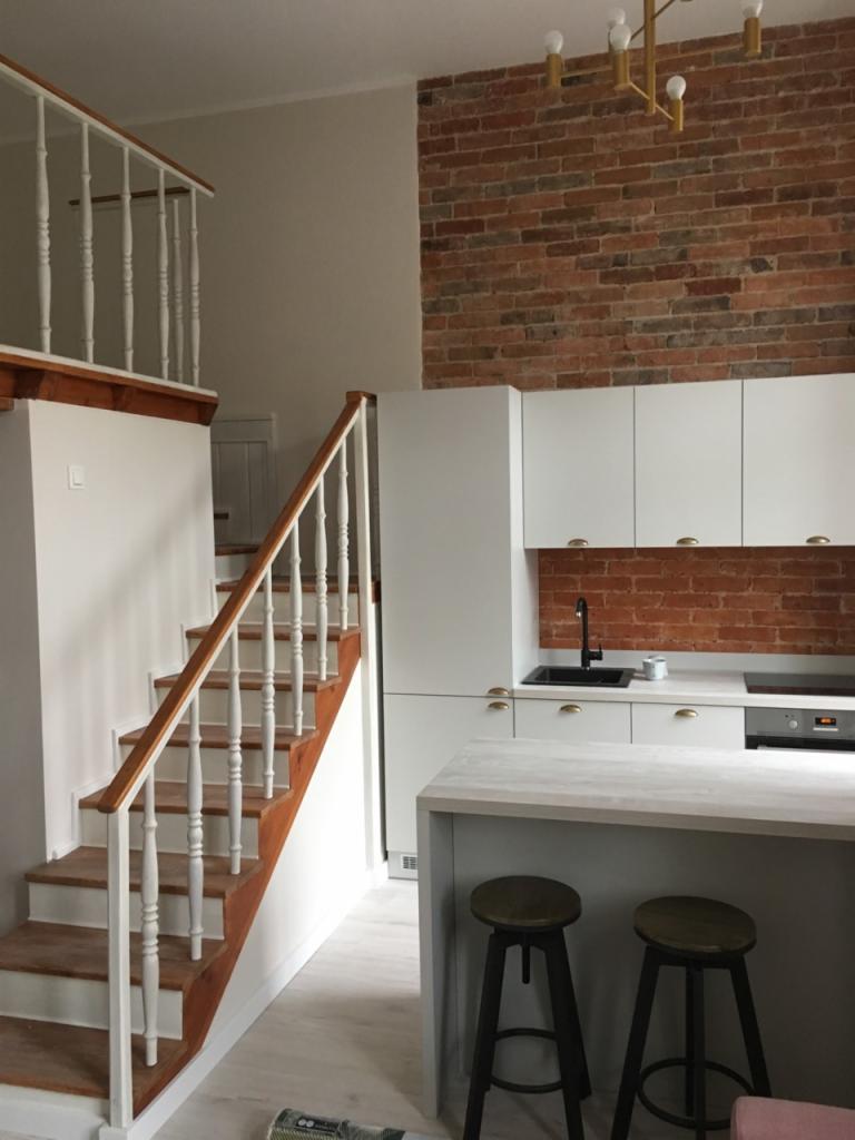 Wnętrze domu z drewnianymi schodami prowadzącymi na antresolę i nowoczesną białą zabudową kuchenną z ceglaną ścianą nad blatem oraz złotym oświetleniem.