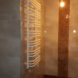 Wnętrze łazienki w trakcie prac wykończeniowych: widoczny grzejnik drabinkowy, płytki w odcieniach beżu i brązu, fragment mozaiki, oraz niedokończone obramowanie lustra.