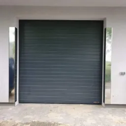 Szara segmentowa brama garażowa z poziomymi przetłoczeniami, zamontowana w białym budynku z dekoracyjnymi, pionowymi, nierdzewnymi elementami po bokach.