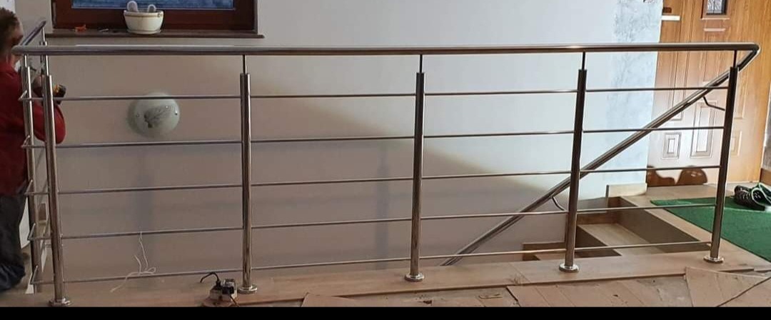 Montaż balustrady nierdzewnej przy schodach wewnętrznych; widoczny pracownik z wiertarką, elementy balustrady z polerowanej stali nierdzewnej oraz schody z drewnianymi stopniami.