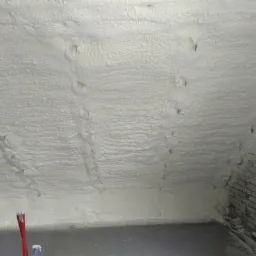 Poddasze ocieplone jasną pianką, widoczne wystające mocowania, fragment ściany z cegły po prawej stronie.