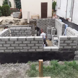 Wznoszenie ścian fundamentowych z bloczków betonowych, zabezpieczonych czarną izolacją, widoczne narzędzia i materiały budowlane, dwóch pracowników w trakcie pracy.