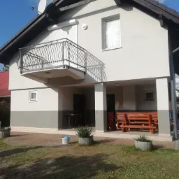 Dwukondygnacyjny dom z balkonem z kutą balustradą, szarą elewacją z ciemniejszym pasem na dole, brązowym dachem i satelitą na dachu, widziany z poziomu trawnika.