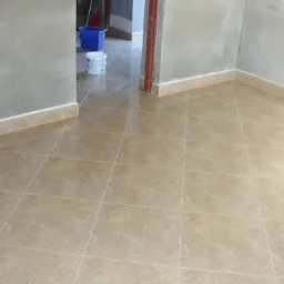 Beżowe płytki podłogowe z delikatnym wzorem marmuru, ułożone w regularną siatkę, z widocznymi fugami i listwami przypodłogowymi w odcieniu zbliżonym do płytek, w tle wiadro i kuweta malarska.