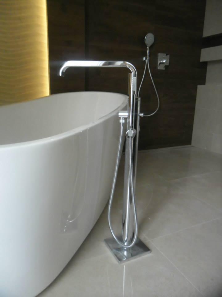 Wolnostojąca bateria wannowa z chromowanym wykończeniem, zamontowana obok białej, owalnej wanny na tle ciemnobrązowej ściany i beżowej podłogi z płytek.