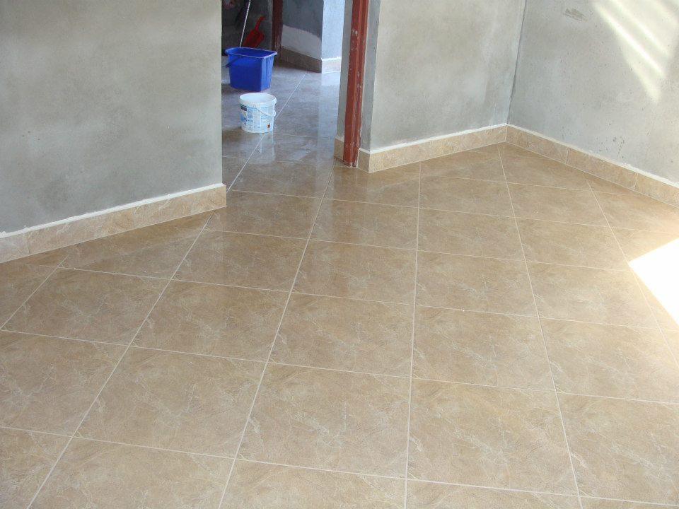 Beżowe płytki podłogowe z delikatnym wzorem marmuru, ułożone w regularną siatkę, z widocznymi fugami i listwami przypodłogowymi w odcieniu zbliżonym do płytek, w tle wiadro i kuweta malarska.