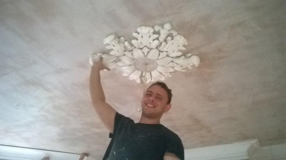 Mężczyzna uśmiechnięty, stojący na drabinie, montuje dekoracyjny element sufitowy w kształcie rozety z liśćmi na nierówno wykończonym suficie.