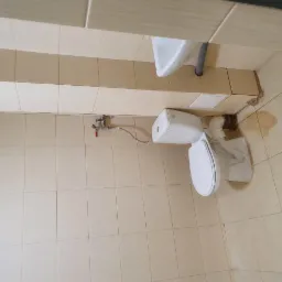 Ukośne ujęcie toalety i umywalki w łazience z beżowymi płytkami. Widoczne ślady po uszczelnianiu przy podłodze i rurach, sugerujące niedawne prace remontowe.