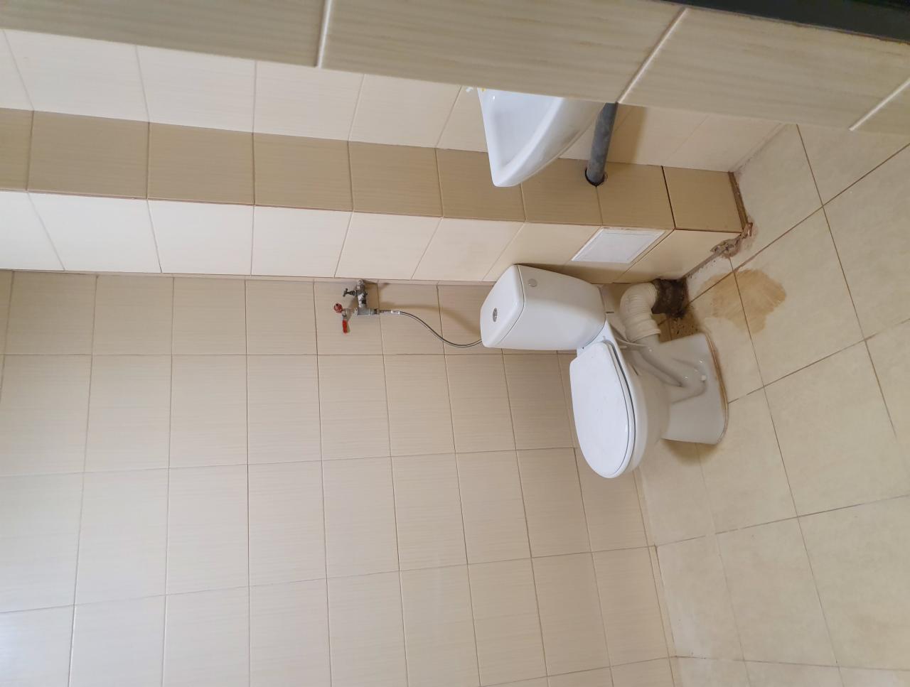 Ukośne ujęcie toalety i umywalki w łazience z beżowymi płytkami. Widoczne ślady po uszczelnianiu przy podłodze i rurach, sugerujące niedawne prace remontowe.