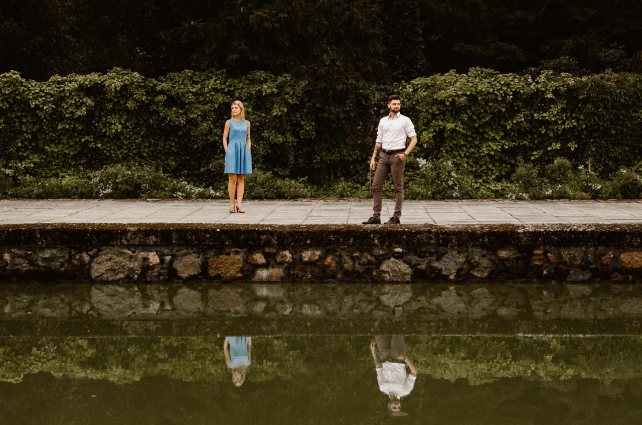Para stoi na kamiennym murku nad wodą z odbiciem, ona w błękitnej sukience, on w białej koszuli i brązowych spodniach, za nimi ściana zieleni.