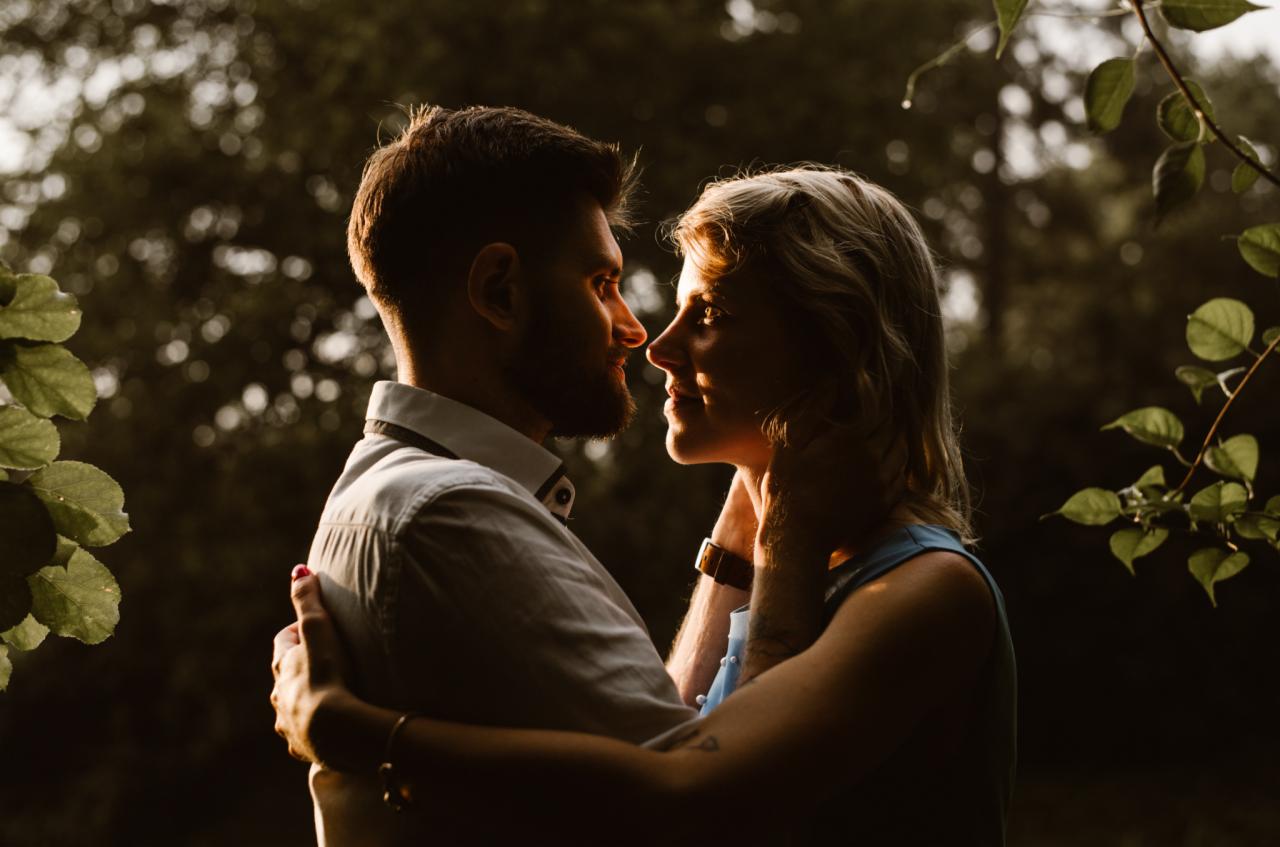 Para zakochanych, mężczyzna z brodą w białej koszuli obejmuje kobietę z blond włosami w plenerze, ciepłe światło zachodzącego słońca.