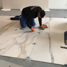 Mężczyzna w pomarańczowych rękawicach układa wielkoformatowe płytki imitujące marmur, używając systemu poziomowania, widoczne narzędzia i akcesoria do układania płytek.