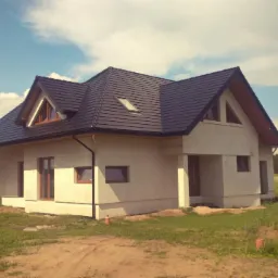 Niedawno wybudowany, parterowy dom z ciemną dachówką i lukarnami, na działce z widocznym nasypem ziemi.