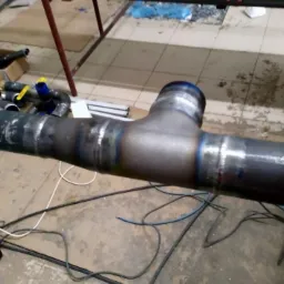 Wspawany trójnik do rury stalowej z widocznymi śladami spawania i elementami instalacyjnymi w tle.