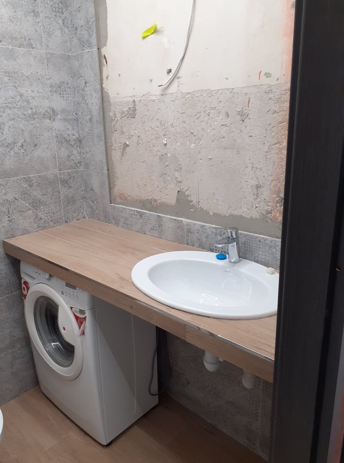 Łazienka w trakcie remontu: umywalka nablatowa na drewnianym blacie zamontowanym nad pralką, ściany częściowo wykończone szarą płytką wzorzystą, fragment ściany z widocznym tynkiem i instalacją.