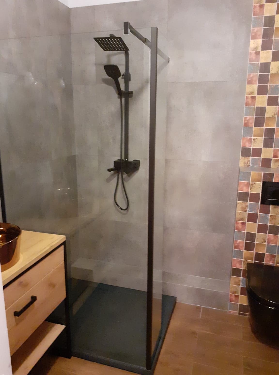 Nowoczesna kabina prysznicowa z czarną armaturą i szklanymi ścianami, widoczna umywalka z drewnianą szafką i fragment toalety z mozaiką w tle.