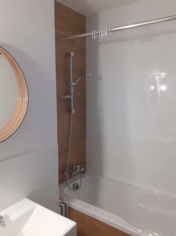 Nowoczesna łazienka z wanną, panelem dekoracyjnym imitującym drewno, okrągłym lustrem i zestawem prysznicowym z deszczownicą.