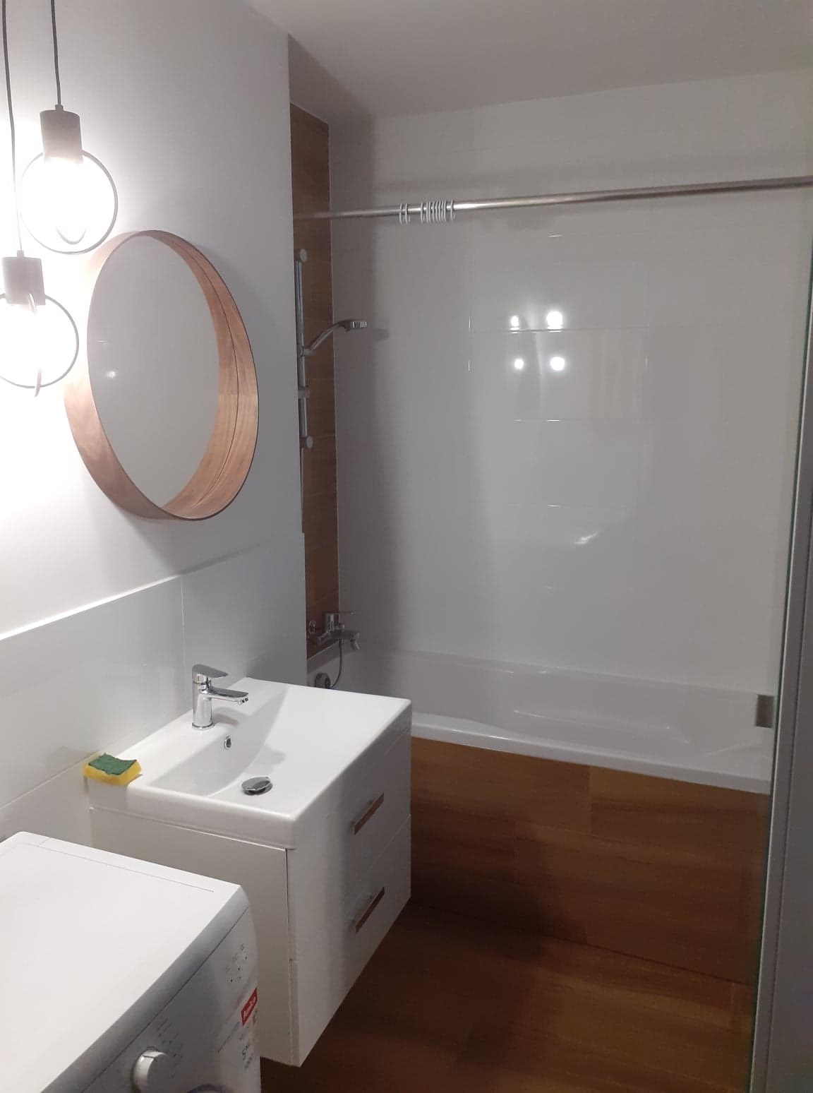 Nowoczesna łazienka z drewnianymi akcentami: okrągłe lustro, wiszące lampy, biała umywalka z szafką, pralka oraz wanna za szklanym panelem.