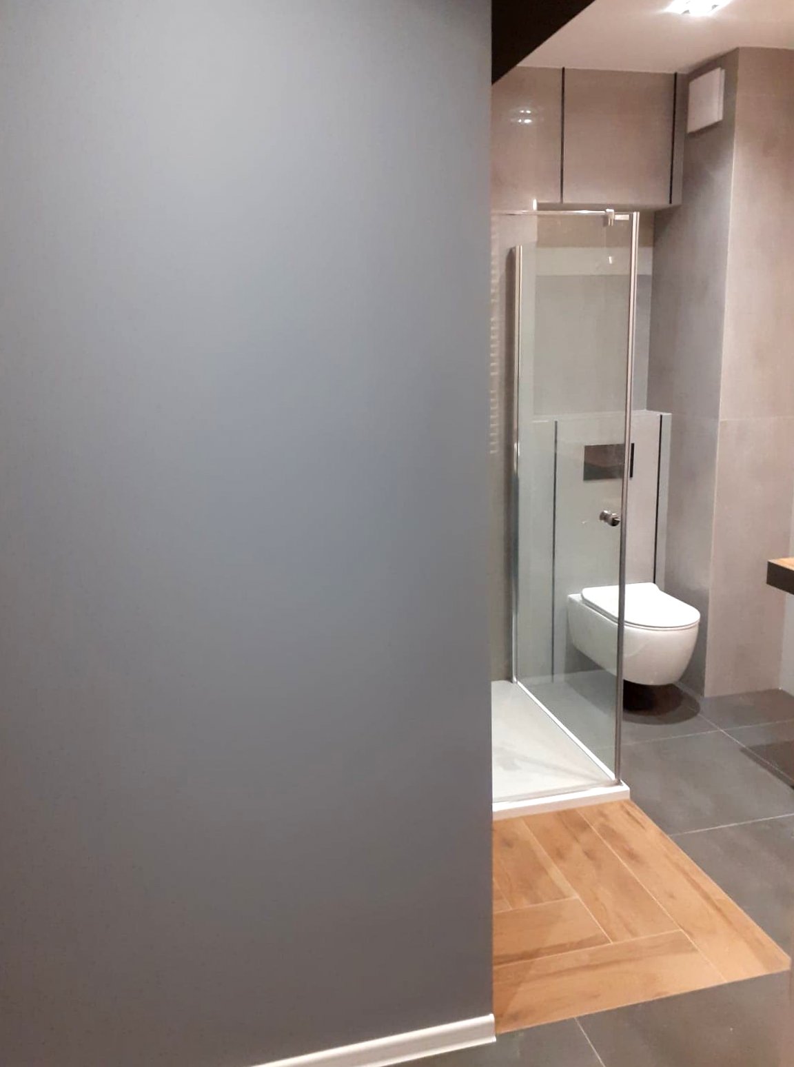 Nowoczesna łazienka z szarą ścianą, kabiną prysznicową ze szklanymi drzwiami, podwieszaną toaletą i fragmentem drewnianej podłogi.