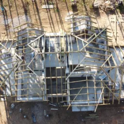 Widok z drona na budowę domu w lesie, widoczne fundamenty i drewniana konstrukcja ścian, obok stoi żółty dźwig i biały samochód dostawczy.