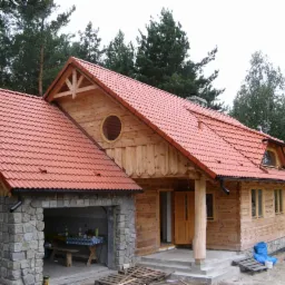 Wykończony drewniany dom z czerwoną dachówką, okrągłym oknem i kamiennym garażem, otoczony drzewami.