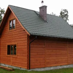 Zewnętrzny widok domu z bali z zielonym dachem z blachy falistej i ceglanym kominem, widoczne okna i weranda z drewnianą balustradą.